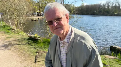 LDRS A man with grey hair and glasses and wearing a beige-coloured coat and a checked shirt is smiling as he stands next to a lake.