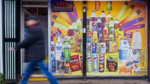 A man walks in front of a convenience store window with product advertising posters on it