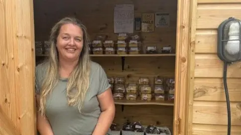 Emma Burton stands in the doorway of the Capel Cake Shed. Shelves behind her are filled with packaged cakes and traybakes.