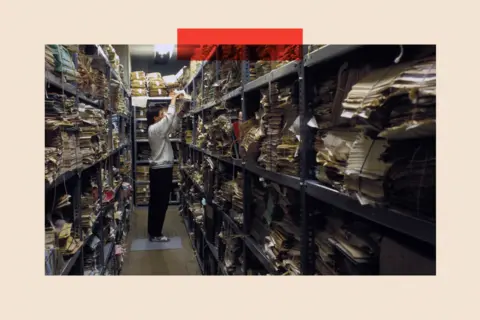 Gamma-Rapho via Getty Images A man in a light grey sweatshirt pulls a stack of files off a shelf in an archive room, which is filled with other files