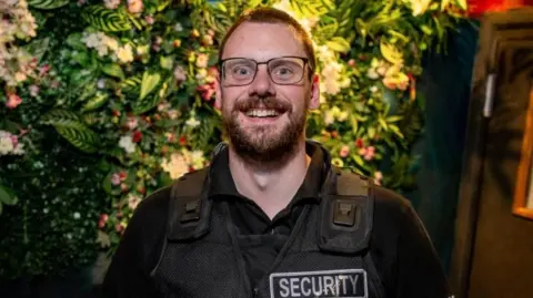 David Barr David Barr in a stab vest that has a label saying security on it. He has a black t-shirt underneath. 