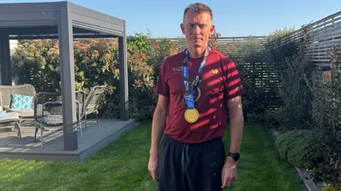 Danny Stapleton in his garden with a medal round his neck, dressed in a red t-shirt and black shorts