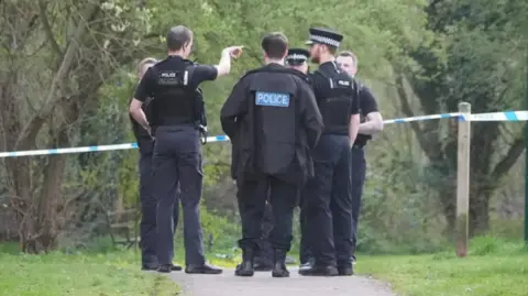 Ant Saddington/BBC A group of police officers wearing black uniforms and hats standing next to some police tape in a wood. One is pointing towards the right.They are standing on a path which has grass on either side. There are trees beyond them.