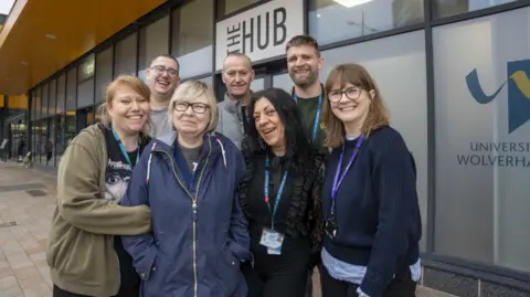City of Wolverhampton Council Four women at the front and three men behind them are posing for the camera. One woman has black hair and another blonde hair and the other women have brown hair. They are standing in front of a building, with the words university and Wolverhampton partly visible on the right.
