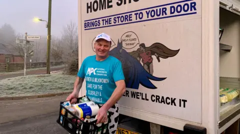 Mr Fowler holds a crate of milk and groceries in front of his delivery van