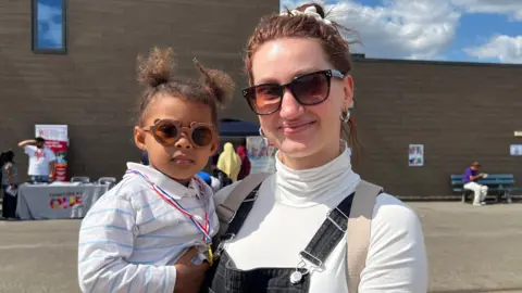 Yana is holding her daughter Maria. Maria has a gold medal over her neck and wearing white shirt with blue stripes and Yana is wearing a black denim dungarees. 
