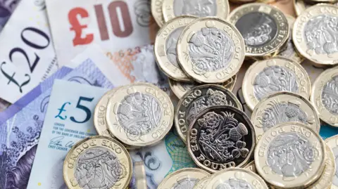 UK Sterling £5, £10 and £20 notes lie on top of one another with a handful of golf and silver pound coins lying on top of the notes.
