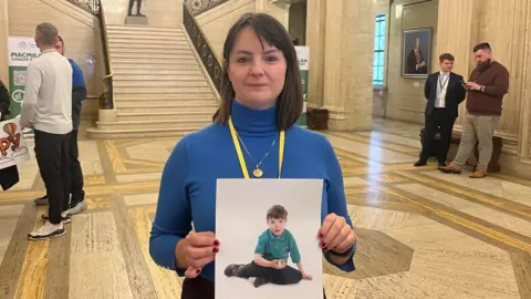 Caroline Devenney stands in the middle of Stormont Great Hall holding a picture of her young son, Harry. She has shoulder-length dark hair and is wearing a yellow lanyard about her neck. She also has a gold necklace. She is looking at the camera with a neutral expression.