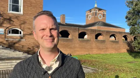 Richard Jefferies, lead member for children's services, wearing a pale yellow shirt and a grey cable-knit quarter-length zipped sweater and standing in front of County Hall offices