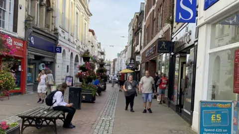 People walking down Barnstaple High Street with shops either side of the road and a person in a white shirt and black trousers sitting on a bench in the foreground.