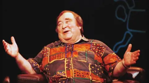 Bernard Manning, who has light brown hair and is wearing an orange and purple short-sleeved shirt in a block pattern, sits in a leather armchair in mid conversation with his arms outstretched.