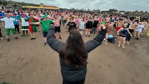 Alan Hughes. Una multitud en la arena, con los brazos extendidos, de pie frente a una mujer de cabello largo y castaño, de espaldas a la cámara. Entre la multitud hay personas vestidas de elfos, con gorros de Papá Noel y trajes de baño.