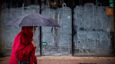 A woman walks by a boarded up UK shop on a rainy day holding an umbrella