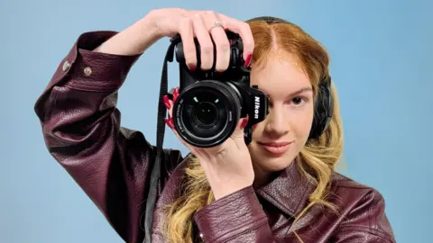 BBC Girl wearing a maroon leather jacket is holding a black Nikon Z8 camera with a large lens and visible lens markings. The camera has a black strap hanging down and is positioned close to the person’s head. Her hand, with long red nails and a silver ring, is gripping the top of the camera
