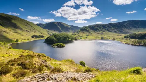 Aerial view of Haweswater. The reservoir is ringed by mountains, green and grassy in the sunlight, which is also reflected on the surface of the water. A promontory extending into the water is covered with conifers, and there are further patches of conifers to either side.
