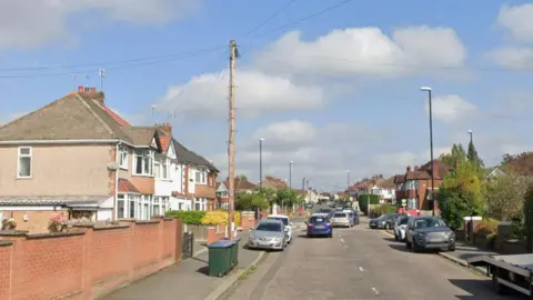 William Bristow Road has houses on both sides of the street and parked cars. There are bins, trees and lamp posts and the junction with Daventry Road is ahead.
