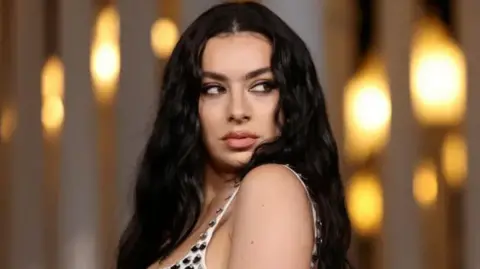 Charli XCX looking off to the side, standing in front of a background featuring glowing lights. She has long, dark hair.