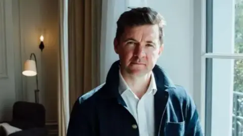 William Layzell has short dark hair and is wearing a dark jacket. He is standing by a window.