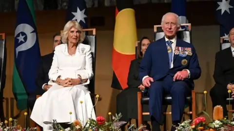 Reuters King Charles and Queen Camilla sat in wooden chairs on a stage with flowers at their feet.