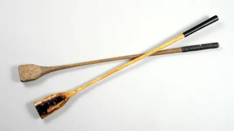 Barnsley Museums A pair of wooden nipsy sticks that are dark in colour with black handles.