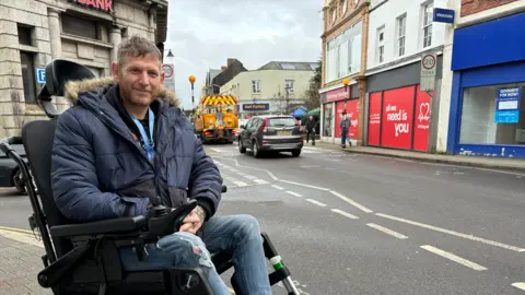 James Ball in a wheelchair wearing a blue parker coat and blue jeans. He is smiling towards the camera. There is a road and buildings behind him.