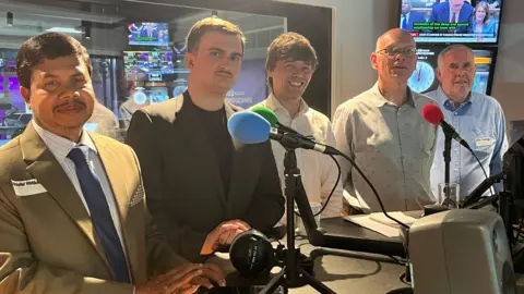 Five smartly-dressed party representatives stand side-by-side in a radio studio.