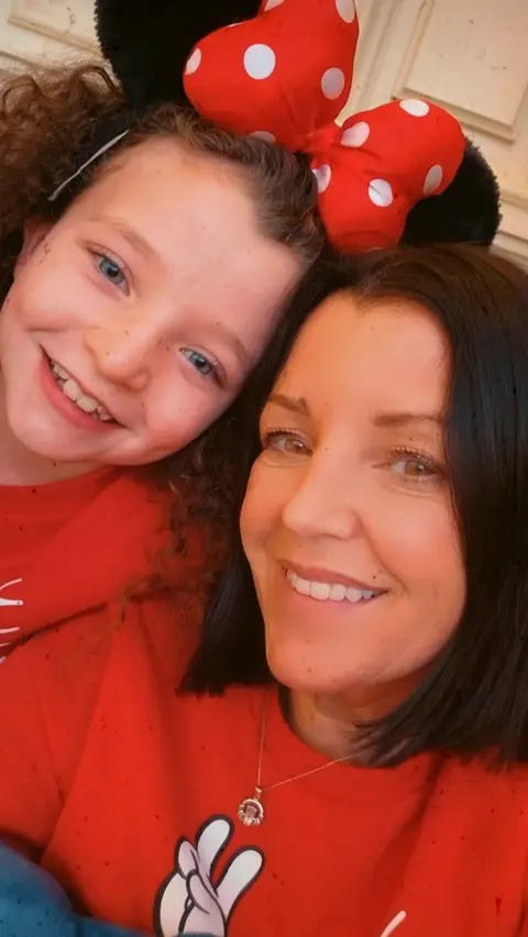 Louise James Rioghnach and Louise pose with their heads close together for a selfie. Rioghnach has curly brown hair and is wearing a red Minnie Mouse style bow with white polka dots on her head. Her mum has a dark brown bob and both are wearing red jumpers.