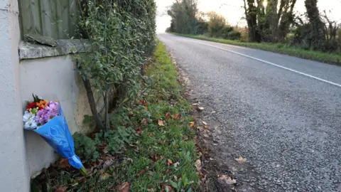 PA Media Flowers wrapped in blue wrapping placed against a wall with fencing by the side of a road.