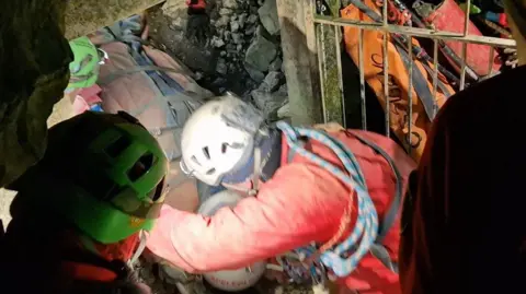 Italian Alpine Rescue Service A woman strapped on to a stretcher, wearing a helmet, is pulled out from a cave in Italy, on the dirt ground beside rocks, through a grate, by rescuers wearing neon clothing, ropes and helmets 