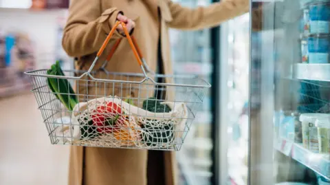 Getty Images Woman in beige coat shopping near freezer section of supermarket adding stuff to basket