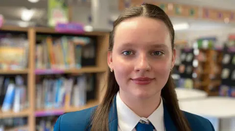 Tetiana is smiling at the camera. She is standing in a library. She has long dark hair and is wearing a school blazer, white shirt and a blue tie. 