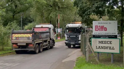 Two large lorries pass each other at the entrance to Meece Landfill, with signs on the right and trees in the background.