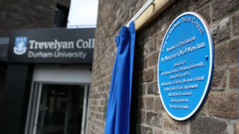 Close-up of a circular blue plaque dedicated to Mo Mowlam. It is attached to a brick wall. Alongside it is a blue curtain that has been pulled to one side.