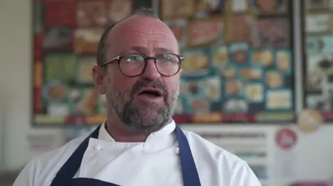 Daniel Smith inside a classroom wearing chef whites and a blue apron. He has a pair of black framed circular glasses on and is looking off camera mid-speech.