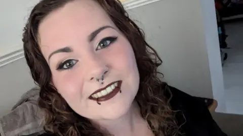 Elizabeth smiles into the camera. She has red lipstick on and piercings in her bottom lip and nose. She has shoulder-length curly brown hair and is wearing a black top.