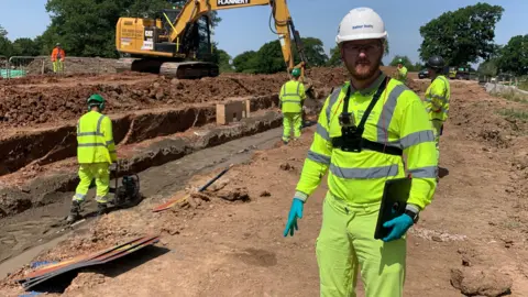 Chris Rodgers pictured at a construction site. He is wearing high vis construction gear and a hard hat. Other workers and a digger can be seen in the background.