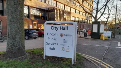 A sign outside a large office building labelled "City Hall." The sign reads "Lincoln Public Services Hub" and lists various public services. There are several parked cars and trees nearby.