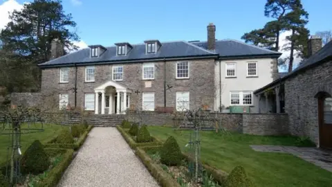 Monmouthshire council A gravel path through a sculpted Victorian garden leading up to the Romanesque entrance, with white pillars of a large stone mansion flanked by ancient trees and a stone barn 