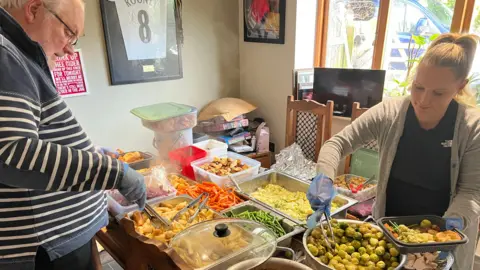 A grey-haired man in a striped jumper is holding some tongs and placing parsnips into a plastic tray. He is standing opposite a woman in a black t-shirt and grey cardigan who is serving up sprouts.