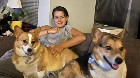 Hannah Cleaver Hannah is smiling towards the camera with two corgis. They are all sitting on a dark grey sofa. Hannah has a light grey blanket on. 