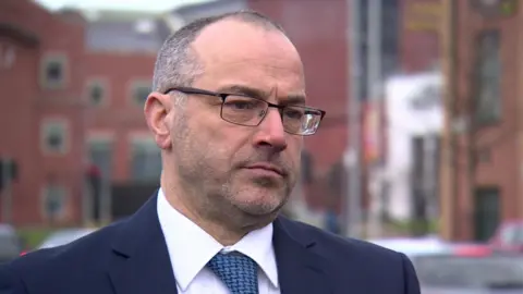 Patrick Corrigan, wearing glasses, with blue jacket, white shirt and blue tie