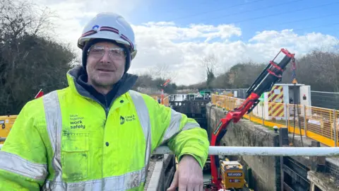 Image shows Stewart Lee standing in front of the canal where the new lock gate is attached to a crane and yet to be lifted. Stewart is slightly to the left and wearing a yellow florescent jacket, with a black fleece underneath. He's wearing a black winter hat with a white helmet on top and is wearing see through protection goggles. Behind him is the red crane arm which is attached by chains to the top of the brand new lock gate which is currently against the canal wall, waiting to be lifted into place. There is metal yellow fencing on the right hand side of the canal wall, above the canal where equipment is being stored.