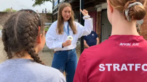 Esme speaking to youngsters at the club while holding her medal. We see the backs of two girls, one with her brown hair in braids, the other with her auburn hair in a bun, looking towards Esme.