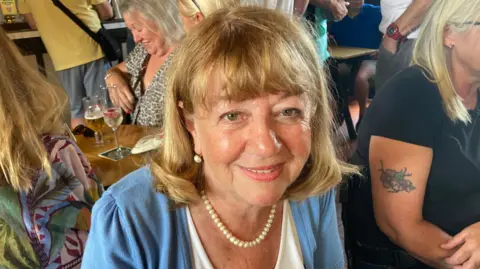 BBC/ Carla Fowler A woman wearing a pearl necklace smiles from the inside of a pub