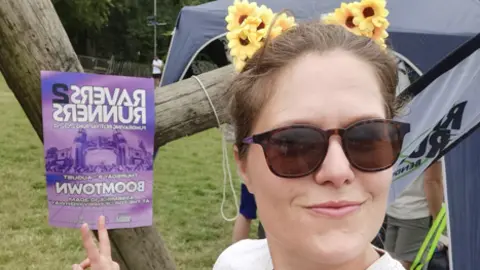 Hannah Ward A close-up of Hannah, who is in her 30s. She is smiling, wearing sunglasses and a hairband featuring bunny ears decorated with tiny sunflowers. She has her two fingers up in a peace sign, and is gesturing towards a Ravers2Runners poster behind her in the field where she is standing