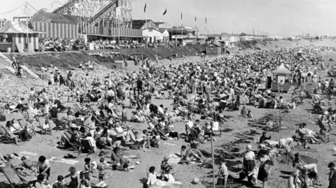 Getty Images A black and white picture of a very crowded beach with funfair rides in the background.