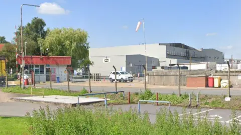 The McCain factory in Easton, near Grantham. A wire fence and small security cabin, with white walls and a red roof, guard a large light grey manufacturing building with a black, white and yellow McCain logo on the side.