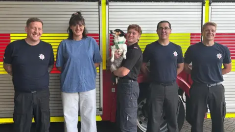 Corby Fire Station Four firefighters, one in the middle, holding a dog, they are all wearing dark T-shirts and trousers. A woman, second from the left, has shoulder length dark hair, white trousers and a blue top. They are all standing in front of a fire engine.