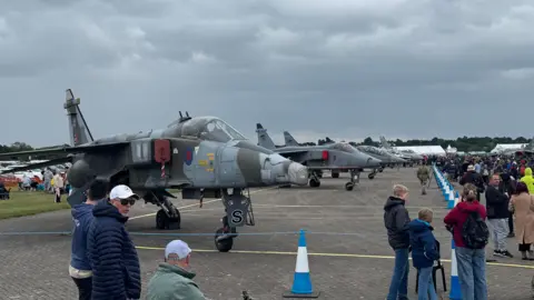 A number of fighter jets lined up in a taped off area on an airfield. People have gathered around to look at the aircraft.