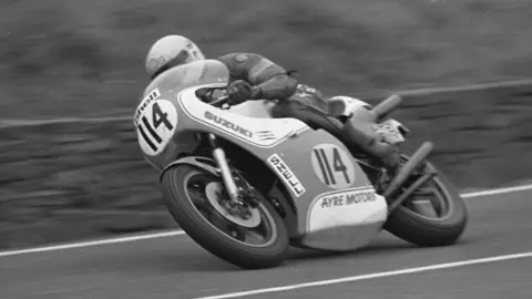 A black and white image of Allan Brew racing on a motorbike. He is leaning down to his right as he turns a corner.